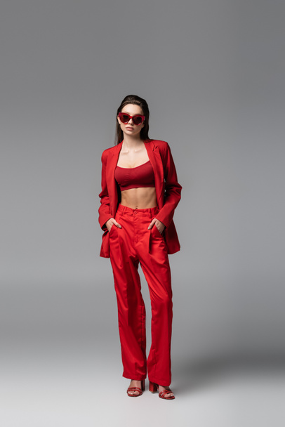 full length of young woman in trendy red suit and sunglasses standing with hands in pockets on dark grey - Fotografie, Obrázek