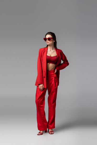full length of young woman in trendy red suit and sunglasses standing with hand on hip on dark grey - Фото, изображение