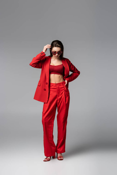 full length of young model in red suit and sunglasses standing with hand on hip on dark grey - Fotó, kép