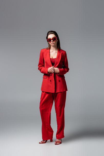 full length of young woman in trendy red suit and sunglasses standing on dark grey - Valokuva, kuva