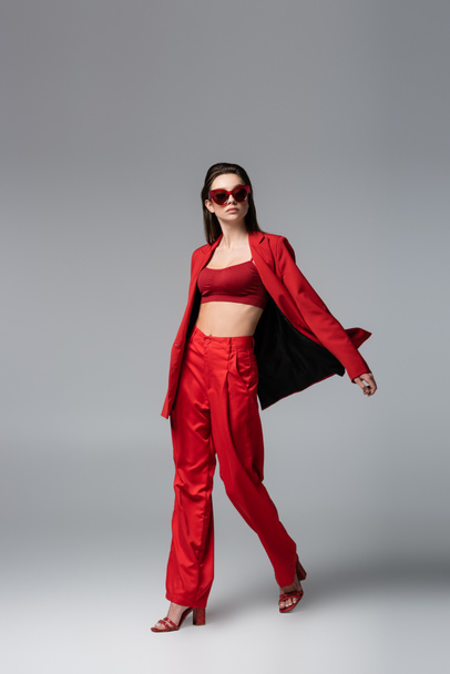 full length of young woman in trendy red suit and sunglasses walking on dark grey - Foto, Imagem