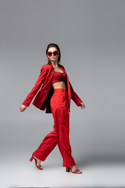 full length of brunette woman in red suit and trendy sunglasses walking on dark grey - Photo, Image