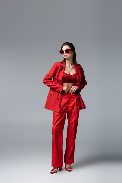 full length of young woman in trendy red suit and sunglasses posing on dark grey - Фото, зображення