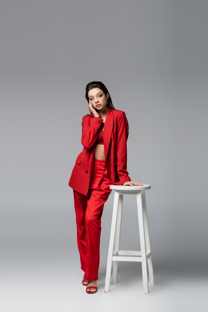 full length of young woman in trendy red suit posing near white chair on dark grey - 写真・画像