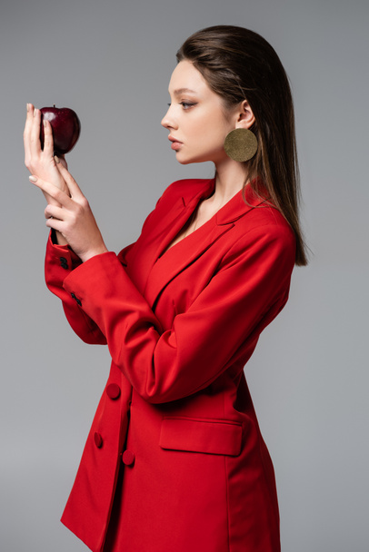 young woman in red suit looking at apple isolated on grey  - Foto, Imagen