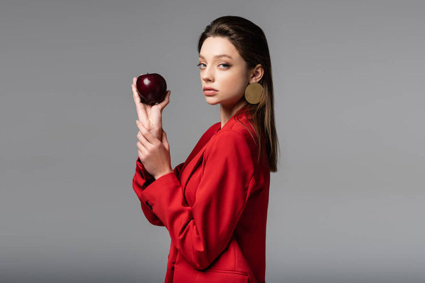 jovem mulher bonita em terno vermelho segurando maçã isolada em cinza  - Foto, Imagem