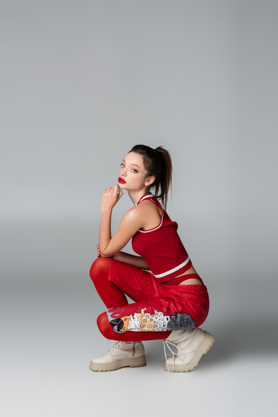 full length of stylish young woman in red sportive outfit and boots sitting while posing on grey - Фото, изображение