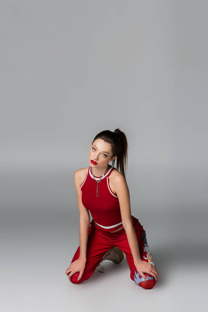 mujer joven de moda en traje deportivo rojo de pie sobre las rodillas en gris - Foto, Imagen