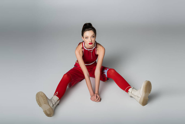 повна довжина стильної молодої жінки в червоному спортивному вбранні та чоботях, що сидять на сірому
 - Фото, зображення