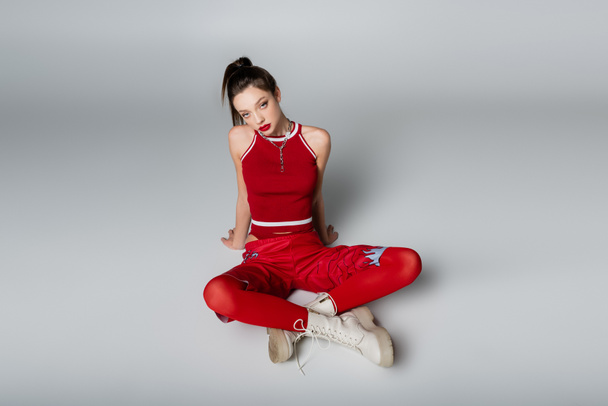 trendiges junges Model in rotem sportlichem Outfit und Stiefeln sitzend mit gekreuzten Beinen auf grau - Foto, Bild