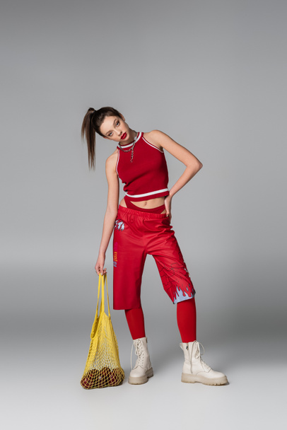 full length of young woman in red sportive outfit holding string bag with apples while posing with hand on hip on grey - Foto, Bild