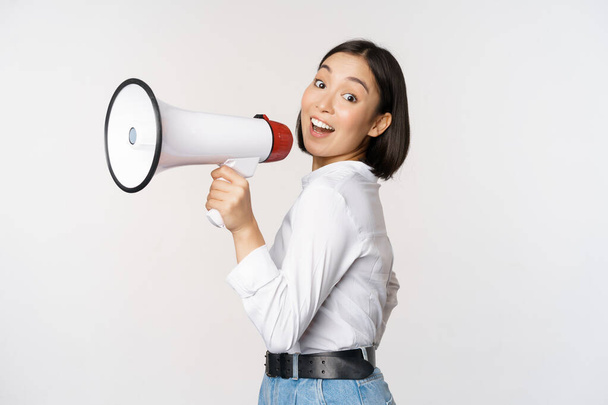 Beautiful young asian woman talking in megaphone, screams in speakerphone and smiling, making announcement, shout out information, standing over white background - Foto, Bild