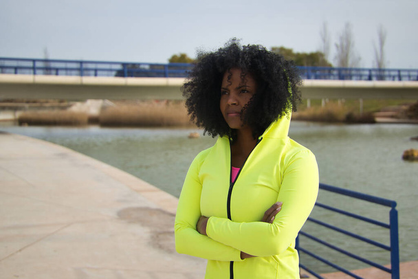 Αφρο-Αμερικανίδα γυναίκα με αφρο μαλλιά και αθλητικά, φοράει φθορίζουσα κίτρινη κουκούλα και κολάν, σταυρωμένα τα χέρια, δείχνει σκεπτική. Fitness έννοια, αθλητισμός, δρόμος, αστικές. - Φωτογραφία, εικόνα