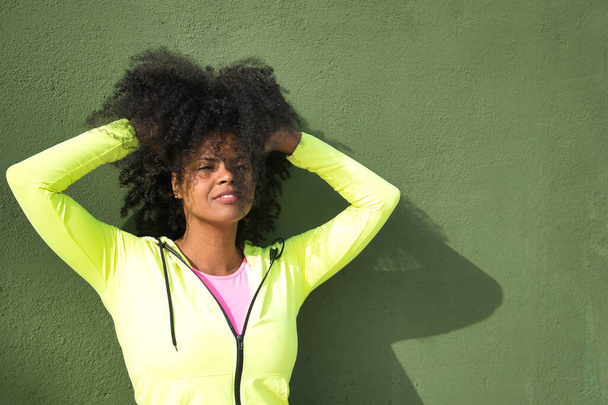 Afrikai-amerikai nő afro hajjal és sportruházattal, fluoreszkáló sárga pulóverben, egy zöld falnak támaszkodva, felemelve a haját, mosolyogva. Fitness koncepció, sport, utca, boldogság, szépség. - Fotó, kép