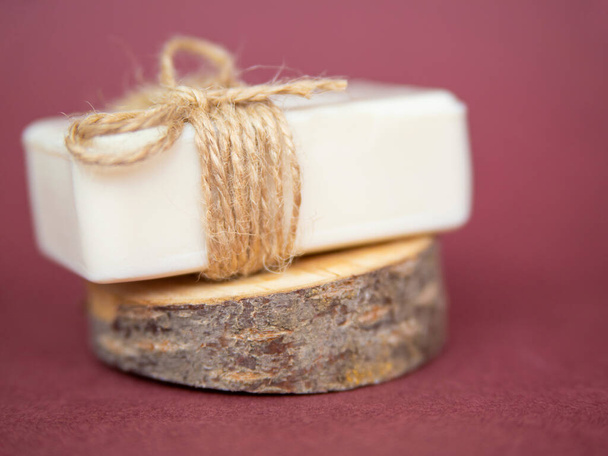 A piece of white soap lies on a cut of a tree on a burgundy background. A natural organic product. Cosmetics for face and body. Environmental friendliness. - Photo, image