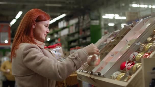 Charmante Shopperin wählt Neujahrsspielzeug, um den Weihnachtsbaum für die Winterferien im Einkaufszentrum zu schmücken, lächelt und blickt in die Kamera - Filmmaterial, Video