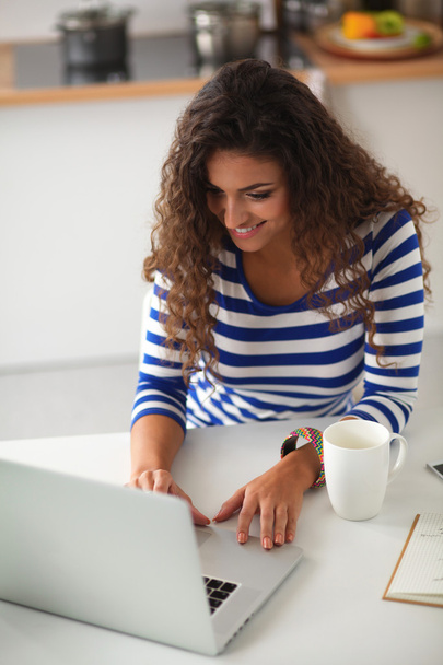 Smiling young woman with coffee cup and laptop in the kitchen at home - 写真・画像