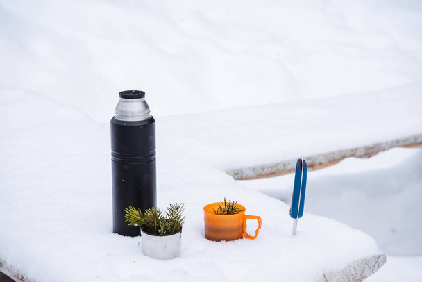 A black thermos and mugs with el's sprig in the snow. A hiking knife will be stuck in the snow. Winter still life in the snow. High quality photo - Foto, Bild