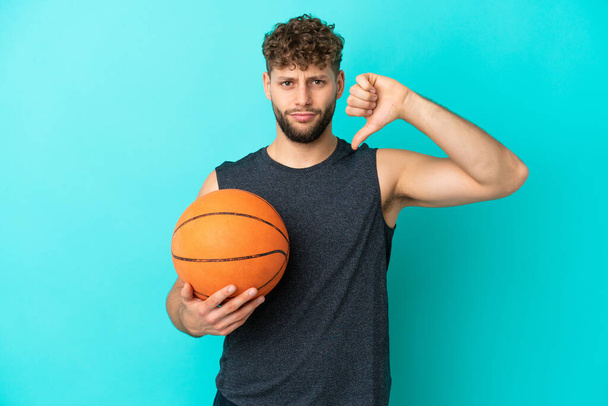 Schöner junger Mann spielt Basketball isoliert auf blauem Hintergrund und zeigt den Daumen nach unten mit negativem Ausdruck - Foto, Bild