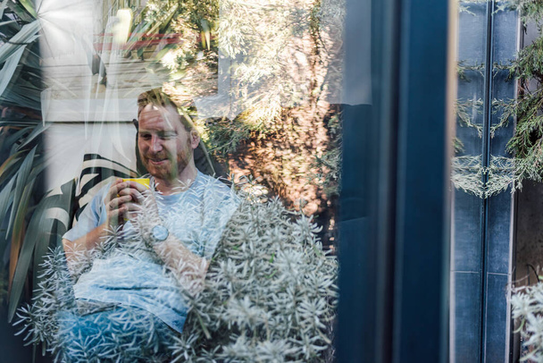 Κοιτάξτε μέσα από το παράθυρο από τον κήπο για χαρούμενα χαμογελαστός νεαρός ενήλικας κάθεται άνετα στο σπίτι σε μεγάλο καναπέ πίνοντας καφέ ή τσάι ξοδεύοντας δωρεάν ελεύθερο χρόνο αναψυχής - Φωτογραφία, εικόνα