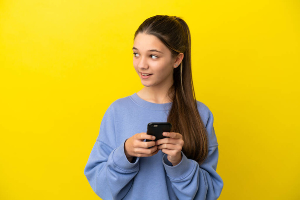 Маленька дівчинка над ізольованим жовтим тлом, використовуючи мобільний телефон і дивлячись вгору
 - Фото, зображення