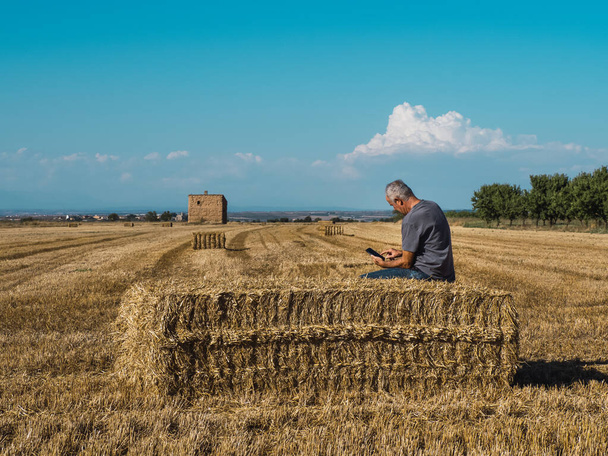 Un granjero mayor sentado en una paca de paja mirando el teléfono móvil en el campo. Concepto agrícola. - Foto, imagen