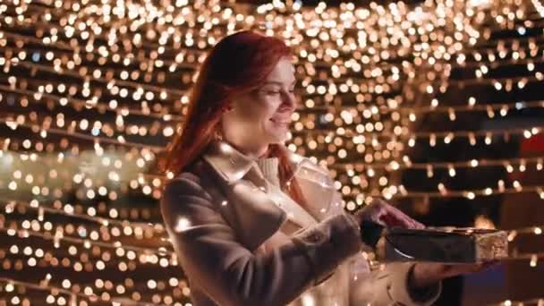 クリスマス・イヴ魅力的な女性が公園の灯りを背景にお祝いの箱を開けてカメラを見て笑っています - 映像、動画