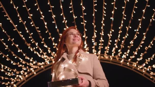 charmante vrouw in bril met cadeau in haar handen wandelingen in prachtig park versierd met een boog van bloemenslingers op kerstavond, glimlachen en kijken naar de camera - Video