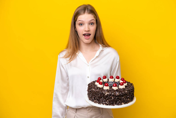 Elinde doğum günü pastası tutan genç Rus kız sarı arka planda şaşırmış bir yüz ifadesiyle izole edilmiş. - Fotoğraf, Görsel