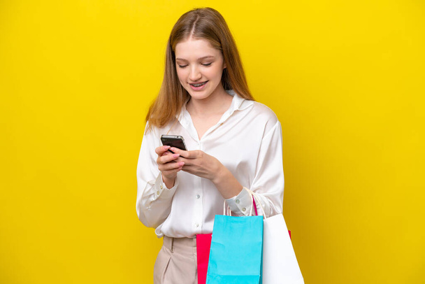 Sarı arka planda izole edilmiş genç bir Rus kız elinde alışveriş çantaları ve bir arkadaşına cep telefonuyla bir mesaj yazıyor. - Fotoğraf, Görsel