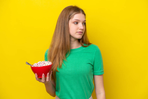 Adolescente russo menina segurando tigela de cereais isolados no fundo amarelo olhando para o lado - Foto, Imagem