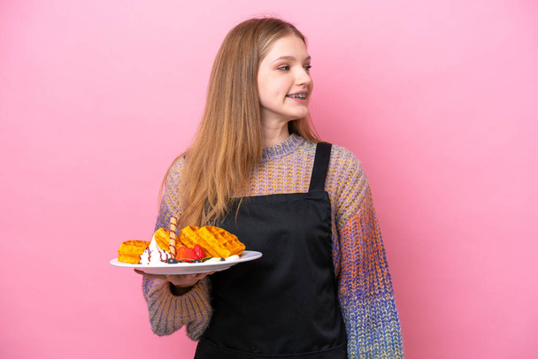 Adolescente rusa chica sosteniendo un waffles aislado en rosa fondo mirando lado - Foto, imagen