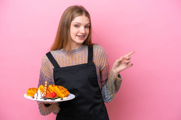Teenager Russian girl holding a waffles isolated on pink background pointing finger to the side - Photo, image