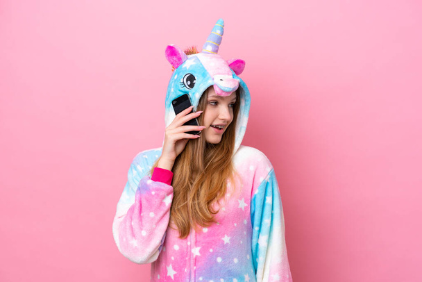 Adolescente chica rusa con pijama de unicornio aislado sobre fondo rosa manteniendo una conversación con el teléfono móvil con alguien - Foto, imagen