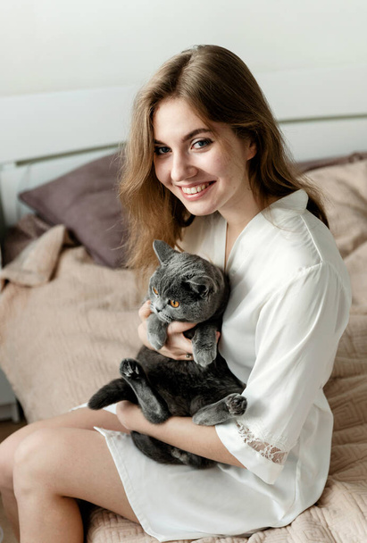 a sexy girl in thin pajamas sits on the bed and holds a beautiful Scottish cat. sexy girl at home in pajamas on a bed with a pet.girl playing with cute cat - Фото, изображение
