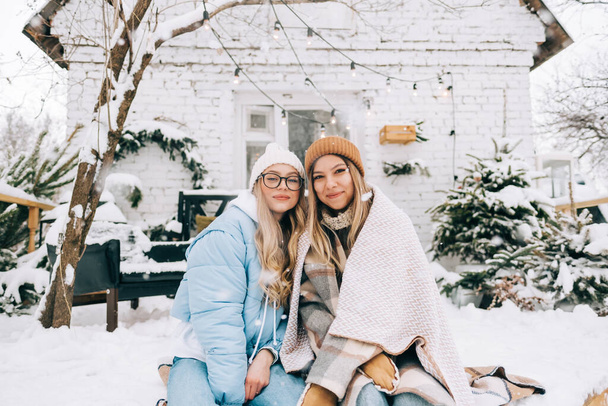 Portrait of two cheerful women friends sitting outdoor on the backyard during snowfall, enjoying winter time. - Photo, Image