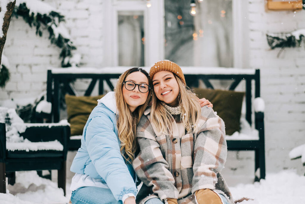 Ritratto di due amiche allegre sedute all'aperto sul cortile dopo la nevicata, godendo l'inverno. - Foto, immagini