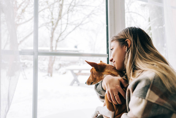 Young caucasian happy woman sitting near window with her dog in winter holidays, looking outside. - Photo, Image