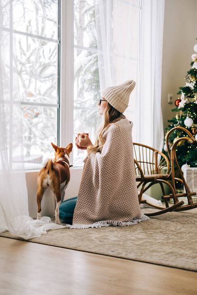 Young caucasian woman sitting near window with her dog and looking outside in the morning, drinking tea and enjoying winter time. - Foto, Imagen