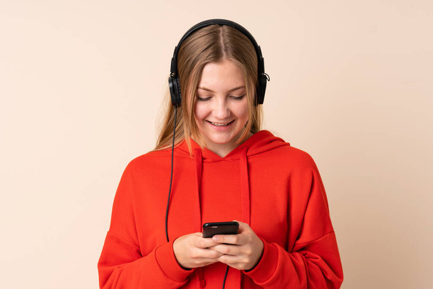 Έφηβη Ουκρανή κοπέλα απομονωμένη σε μπεζ φόντο ακούγοντας μουσική και κοιτώντας στο κινητό - Φωτογραφία, εικόνα