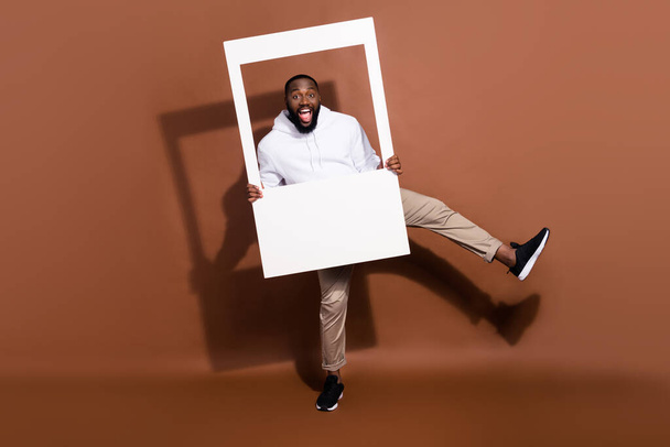 Photo of surprised excited guy wear white hoodie dancing holding paper frame isolated brown color background - Foto, Imagem