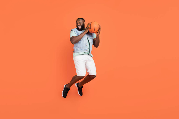 Photo of excited funny afro guy wear denim vest holding ball jumping high isolated orange color background - Fotoğraf, Görsel