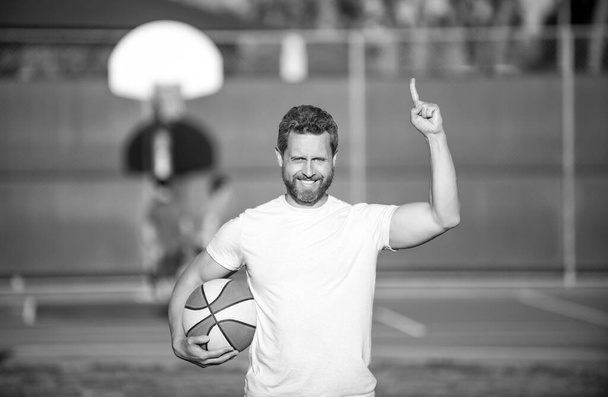 счастливый мускулистый мужчина спортсмен баскетболист поднял палец держать мяч на открытом воздухе, спорт - Фото, изображение