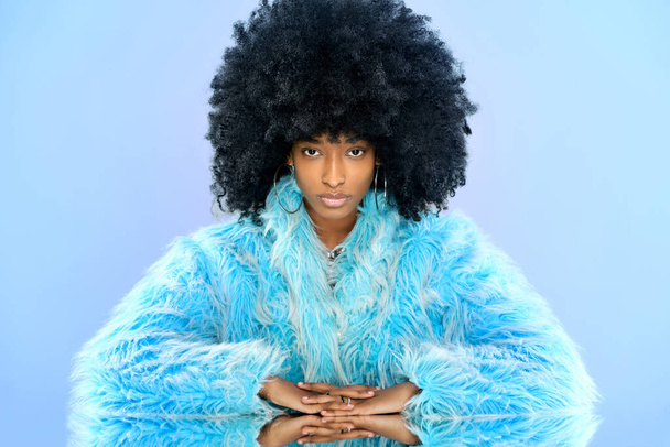 Modelo femenino dominicano con peinado afro en abrigo azul piel mirando a la cámara en la mesa espejada sobre fondo azul - Foto, Imagen