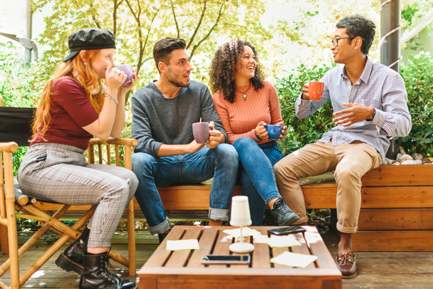 Группа мультикультурных молодых друзей зависает на террасе, расслабляясь за кружками кофе, смеясь и шутя на высоком ключевом фоне - Фото, изображение