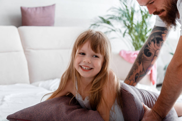 Portrait of child kid girl laughs play with dad. Father with daughter spending time together play in pillow fight in pajamas in bed. Bald bearded Caucasian man in tattoos and tender little daughter. - 写真・画像