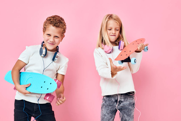 Κομψό μικρό αγόρι και χαριτωμένο κορίτσι με ακουστικά skateboards σε χέρια ροζ χρώμα φόντο - Φωτογραφία, εικόνα