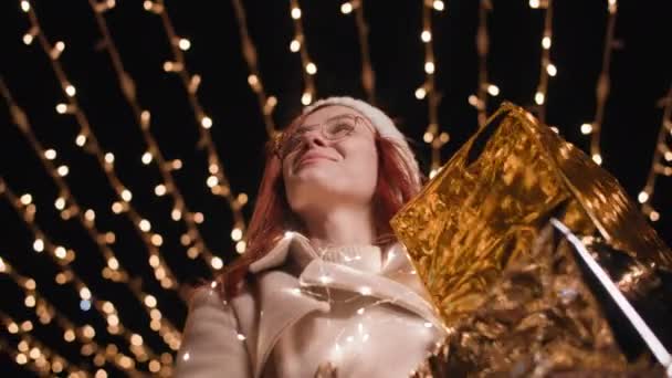 ambiance festive, femme joyeuse avec des cadeaux dans ses mains marche dans l'arc de lumières incandescentes en fin de soirée en plein air - Séquence, vidéo