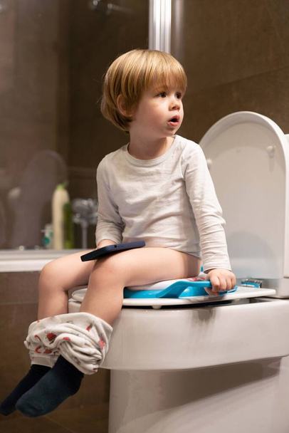 Cute maluch chłopiec, siedzi na toalecie, za pomocą telefonu komórkowego. - Zdjęcie, obraz