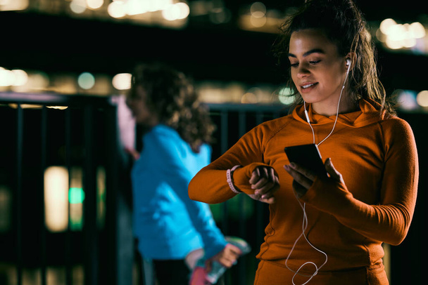 Молода усміхнена нічна бігунка стоїть на відкритому повітрі вночі, використовуючи смартфон і дивлячись на розумний годинник, щоб перевірити частоту серцевих скорочень
. - Фото, зображення